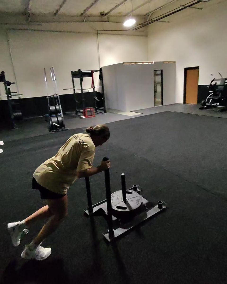 A high-school athlete driving a weighted sled across the gym floor, body angled forward.
