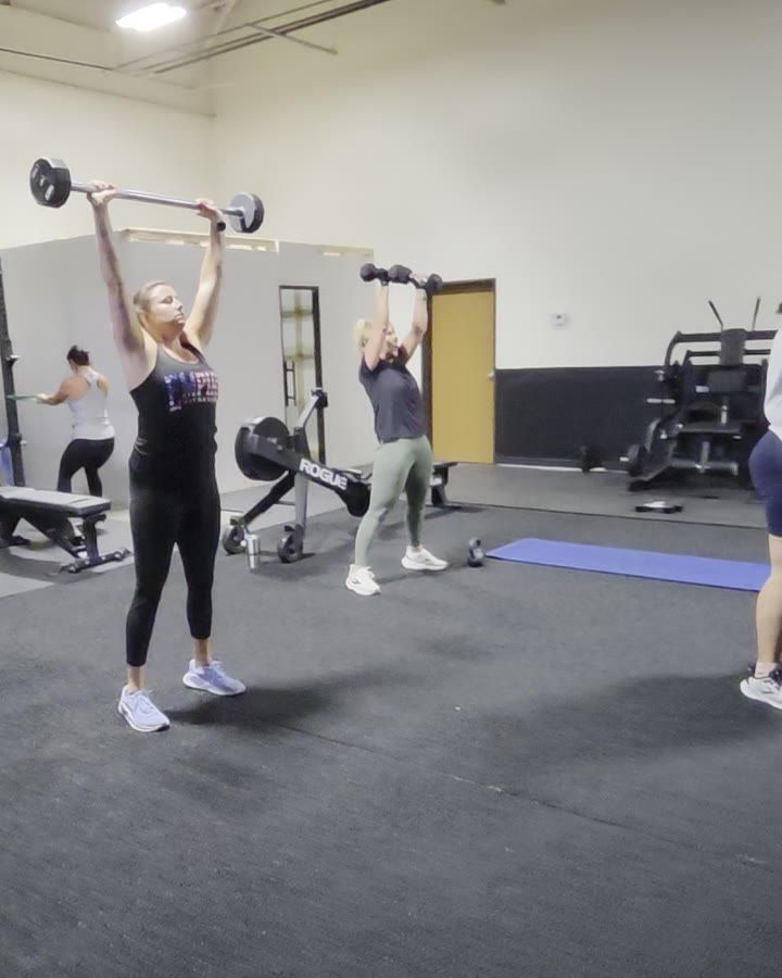 Several women in an Athletic Strength class pressing weights overhead at the same time, one locking out a barbell while the others press dumbbells.