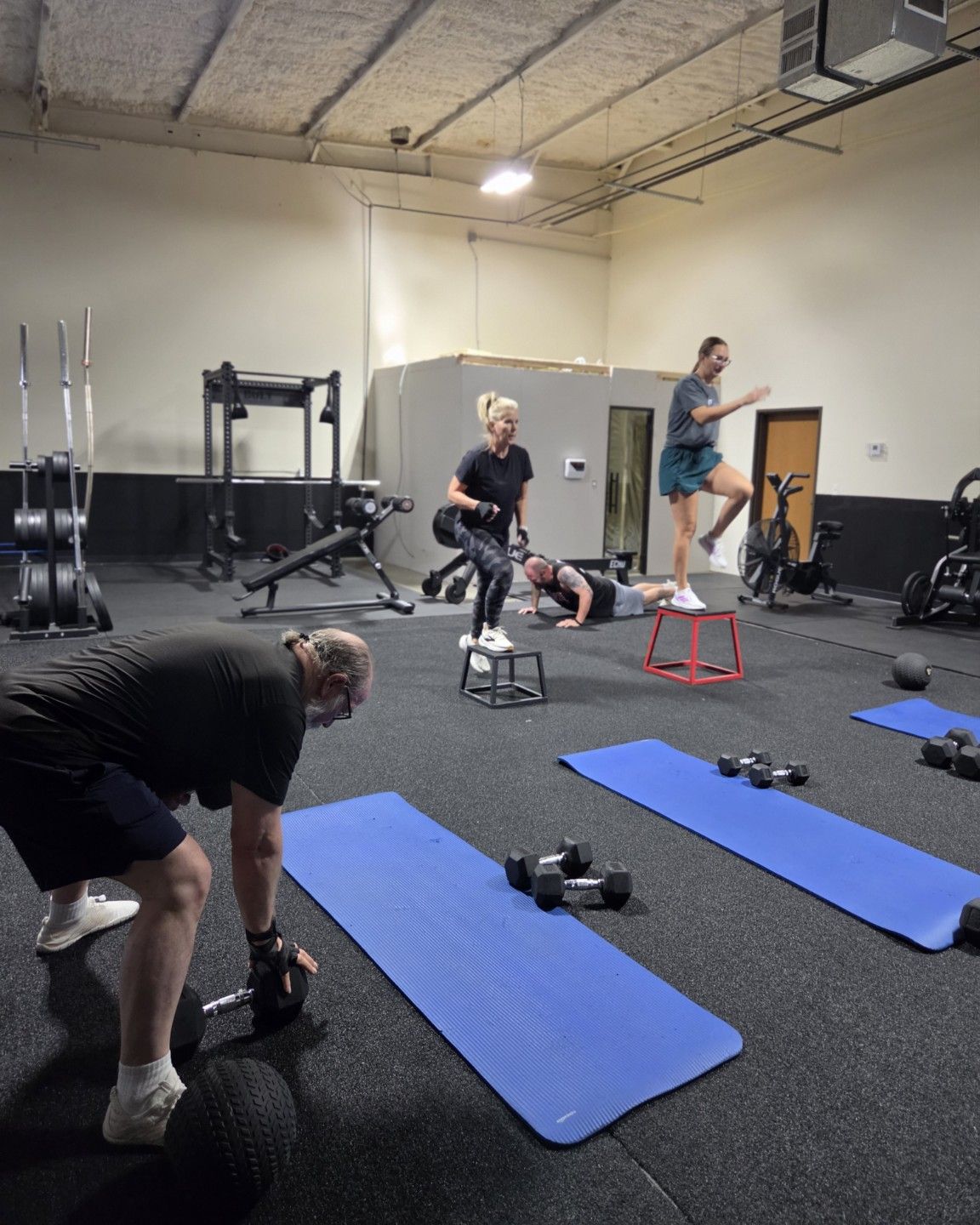 Four adults training at separate stations in an Athletic Strength session at Empire Fitness: one lifting dumbbells from the floor, two stepping onto boxes and one holding a push-up position.