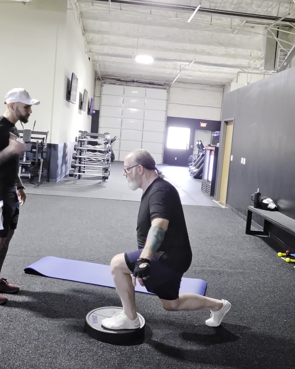 An adult client lowering into a controlled reverse lunge with one foot on a stability dome, while his coach looks on from a few feet away.