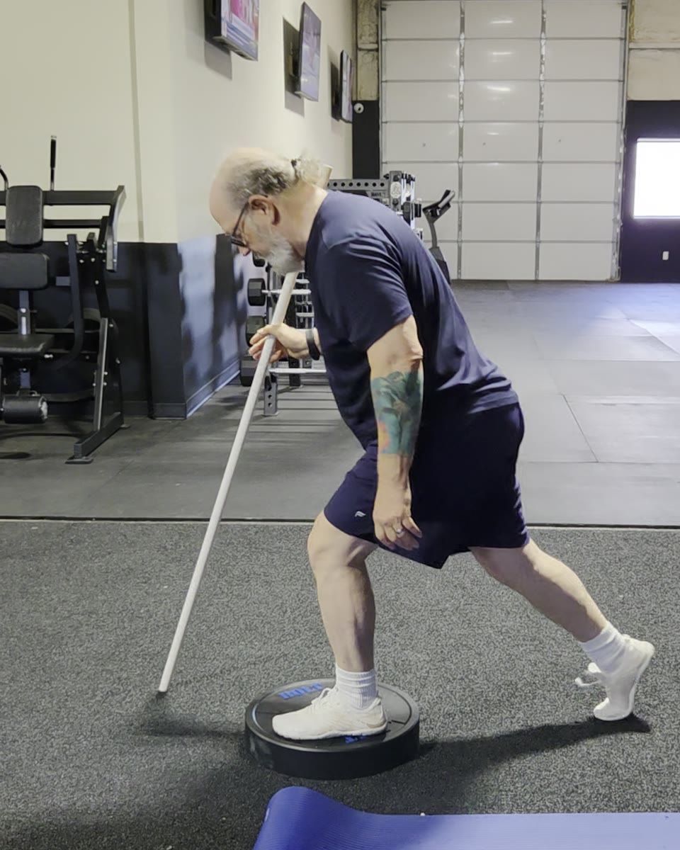 An adult client holding a dowel for balance while lowering into a split squat with one foot on a stability dome.