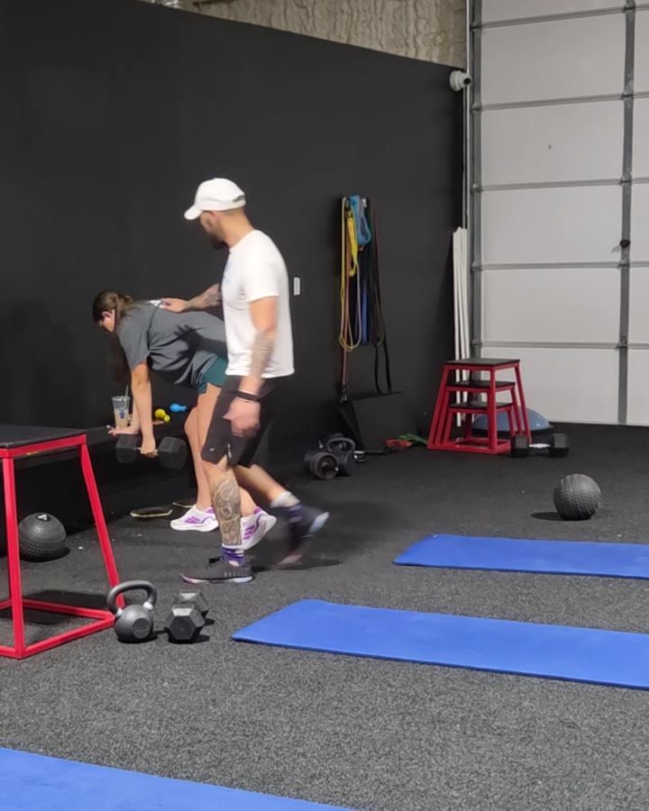 Jonah reaching in to coach a client as she lifts a dumbbell from the floor during a session at Empire Fitness, with weights and mats set out across the training floor.